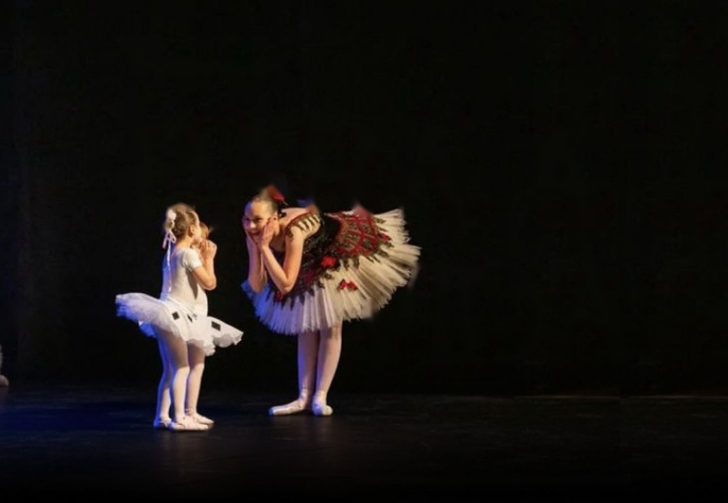 Aluna pequena de tutu branco conversa com uma bailarina mais velha no palco